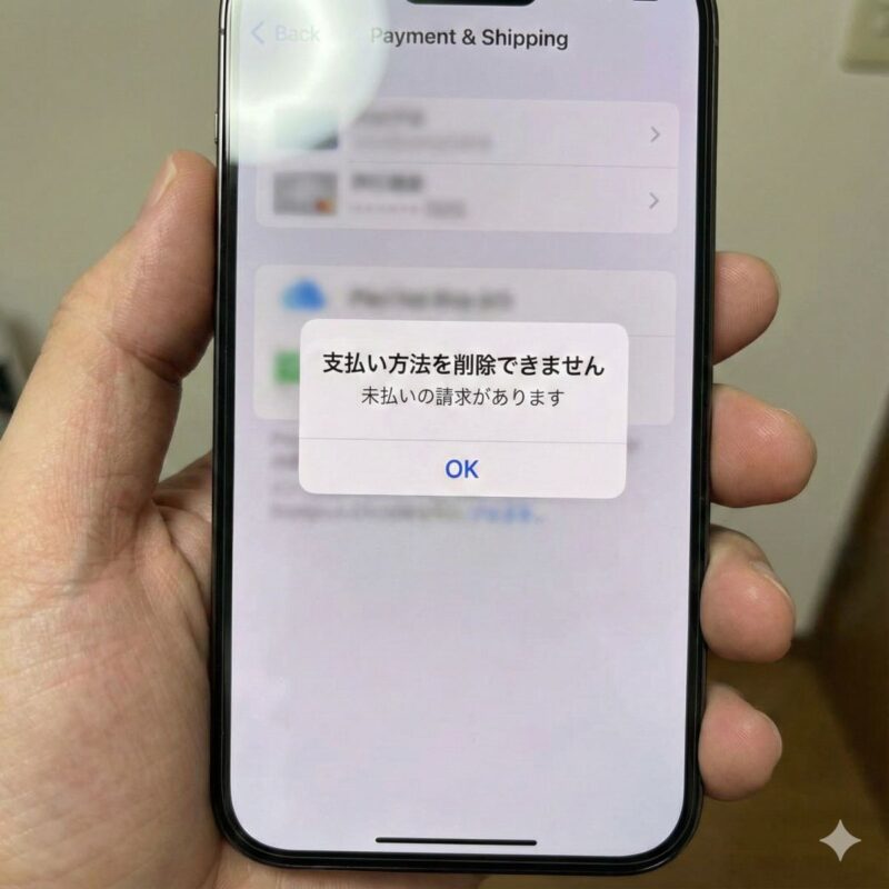 iPhoneの画面中央に、iOSのシステム警告ポップアップが表示されている様子。「支払い方法を削除できません」というタイトルと、「未払いの請求があります」というエラーメッセージ、そして「OK」ボタンが表示されている緊迫した状況。