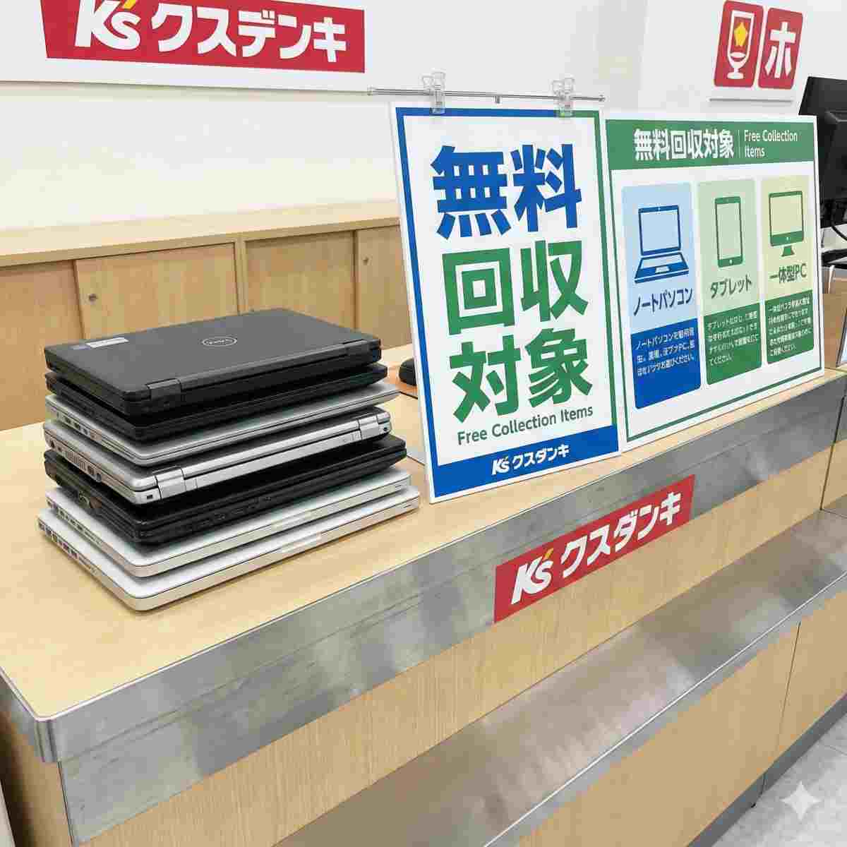 ケーズデンキのブランド名が入った店舗のリサイクルカウンターに整然と積み上げられた様々なノートパソコン。その隣には、「無料回収対象」と書かれた目立つ看板と、対象品目（ノートパソコン、タブレット、一体型PC）のアイコンがリストアップされたインフォグラフィックボードがある。人物はいない。